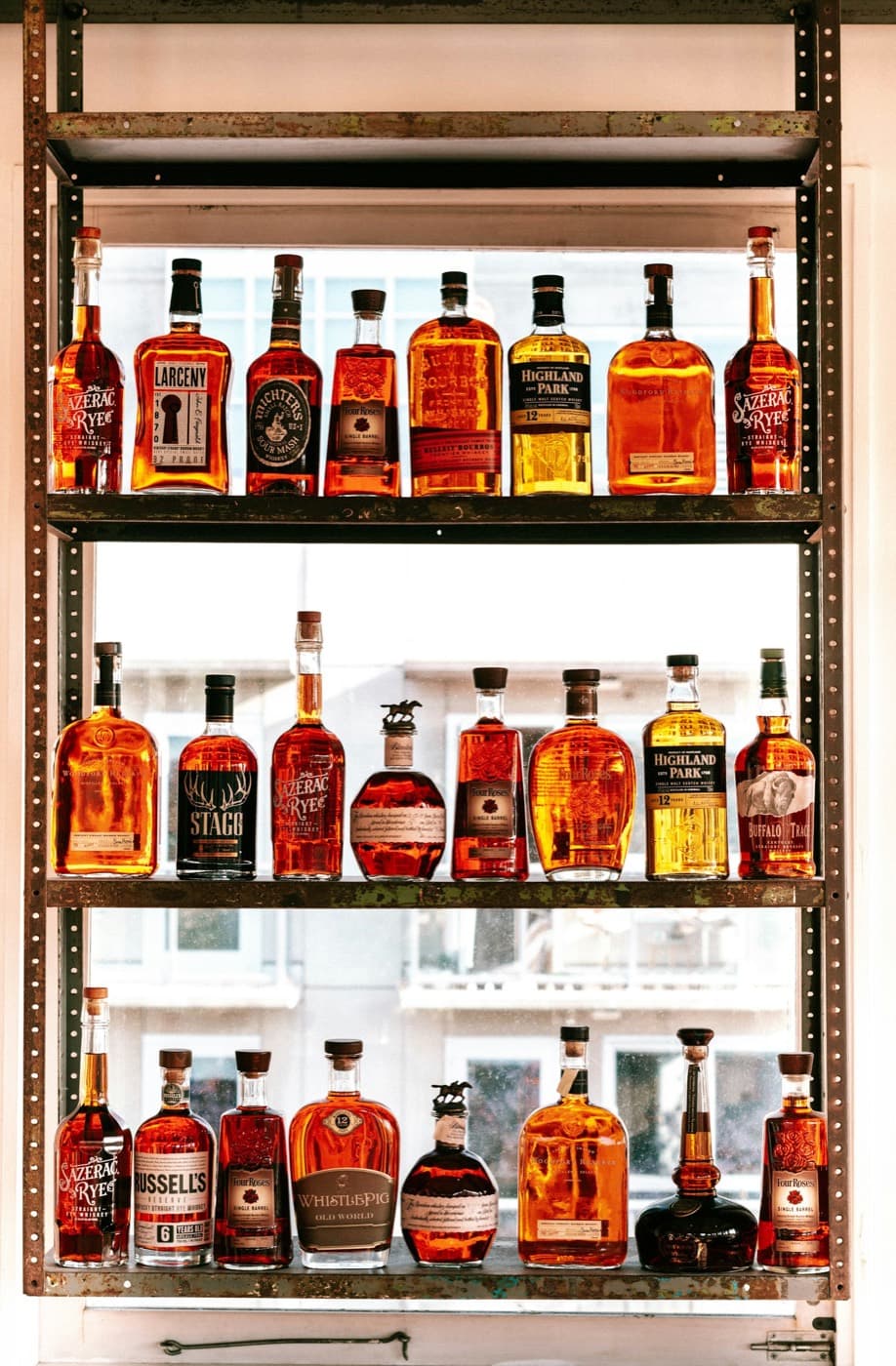 Three steel shelves of bourbon and rye bottles in a bright window